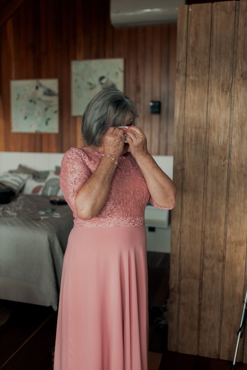 Senior woman in pink lace dress wiping tears, capturing an emotional moment indoors.